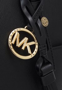 Melna teksturēta ādu soma ar zelta apaļo logo piekariņu, uz kura redzami "MK" un "MICHAEL KORS", ar pīto siksniņu detaļu.