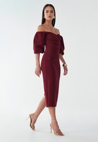 Abito midi bordeaux con spalle scoperte, maniche a palloncino e dettagli arricciati, con uno spacco laterale, abbinato a sandali con tacco alto e cinturini dorati.