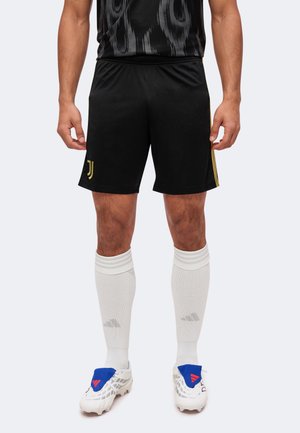 Homme portant un short de football noir avec le logo de la Juventus, des chaussettes blanches montantes et des crampons blancs avec des accents bleus et rouges, debout sur un fond neutre.