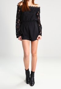Top negro de encaje sin hombros con mangas largas combinado con pantalones cortos negros. El modelo lleva botas negras de tobillo; la tela es suave y con textura.