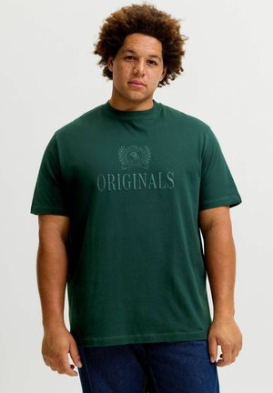 Mand med krøllet hår iført en mørkegrøn t-shirt med teksten "ORIGINALS" og blå jeans, stående foran en ensfarvet lys baggrund.
