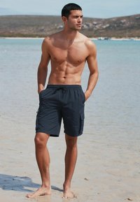 Mornarsko plave kupaće gaće od vodootpornog materijala, s elastičnim pojasom, kravatom i bočnim džepovima, prikazane na pješčanoj plaži.