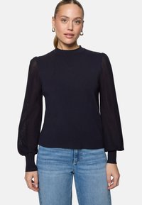 Femme à la peau claire et aux cheveux blonds relevés, portant des boucles d'oreilles anneaux dorées, un pull bleu marine avec des manches bouffantes transparentes et un jean bleu.
