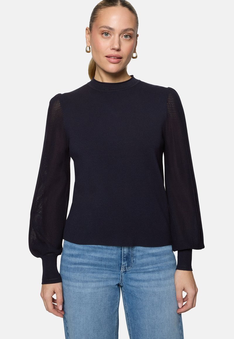 Femme à la peau claire et aux cheveux blonds relevés, portant des boucles d'oreilles anneaux dorées, un pull bleu marine avec des manches bouffantes transparentes et un jean bleu.