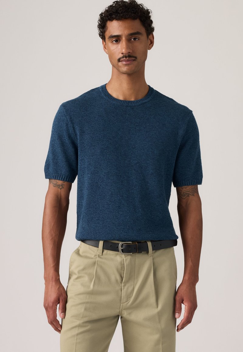 Mann trägt einen blauen kurzärmeligen Strickpullover und beige Hose mit schwarzem Gürtel, steht vor einem schlichten hellen Hintergrund.