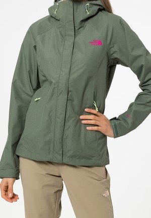 Osoba ubrana w zieloną kurtkę North Face z kapturem i zamykanymi kieszeniami oraz beżowe spodnie North Face, stojąca z jedną ręką na biodrze.