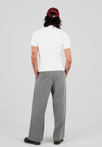 Mężczyzna z ciemnymi włosami, noszący czerwoną czapkę, białą koszulę z krótkim rękawem, szare szerokie spodnie i czarne buty, stojący z rękami w kieszeniach, odwrócony plecami.