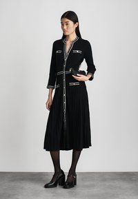 Robe plissée noire avec des bordures blanches et des boutons décoratifs. Manches longues, coupe ajustée, associée à des chaussures noires à talons. Modèle tenant une pochette.