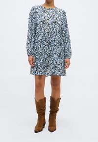 Personne portant une robe à manches longues bleue et blanche à fleurs assortie à des bottes mi-mollet en daim marron, debout devant un fond uni.