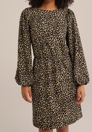 Nainen, joka pitää päällään polvipituisen, pitkähihallisen leo-kuosisen mekon, jossa on rypytetyt hihansuut ja vyötäröltä kiristetty, seisoo yksinkertaisen taustan edessä.