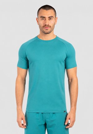 Homme aux cheveux courts et à la barbe taillée, portant une chemise turquoise à manches courtes et un pantalon assorti, debout devant un fond clair uni.