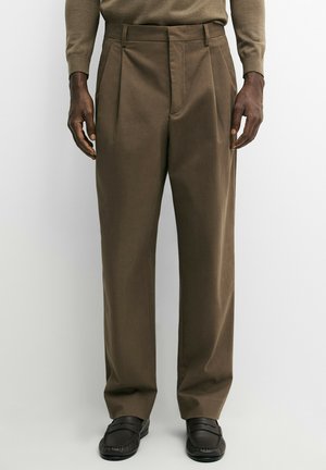 Brune plisserede bukser båret med matchende brun langærmet top og sorte læder loafers på en stående person.
