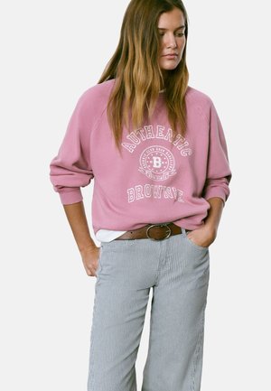 Femme portant un sweat-shirt rose ample avec le texte « Authentic Brownie » et un pantalon rayé, se tenant avec les mains dans les poches, regardant vers le bas.