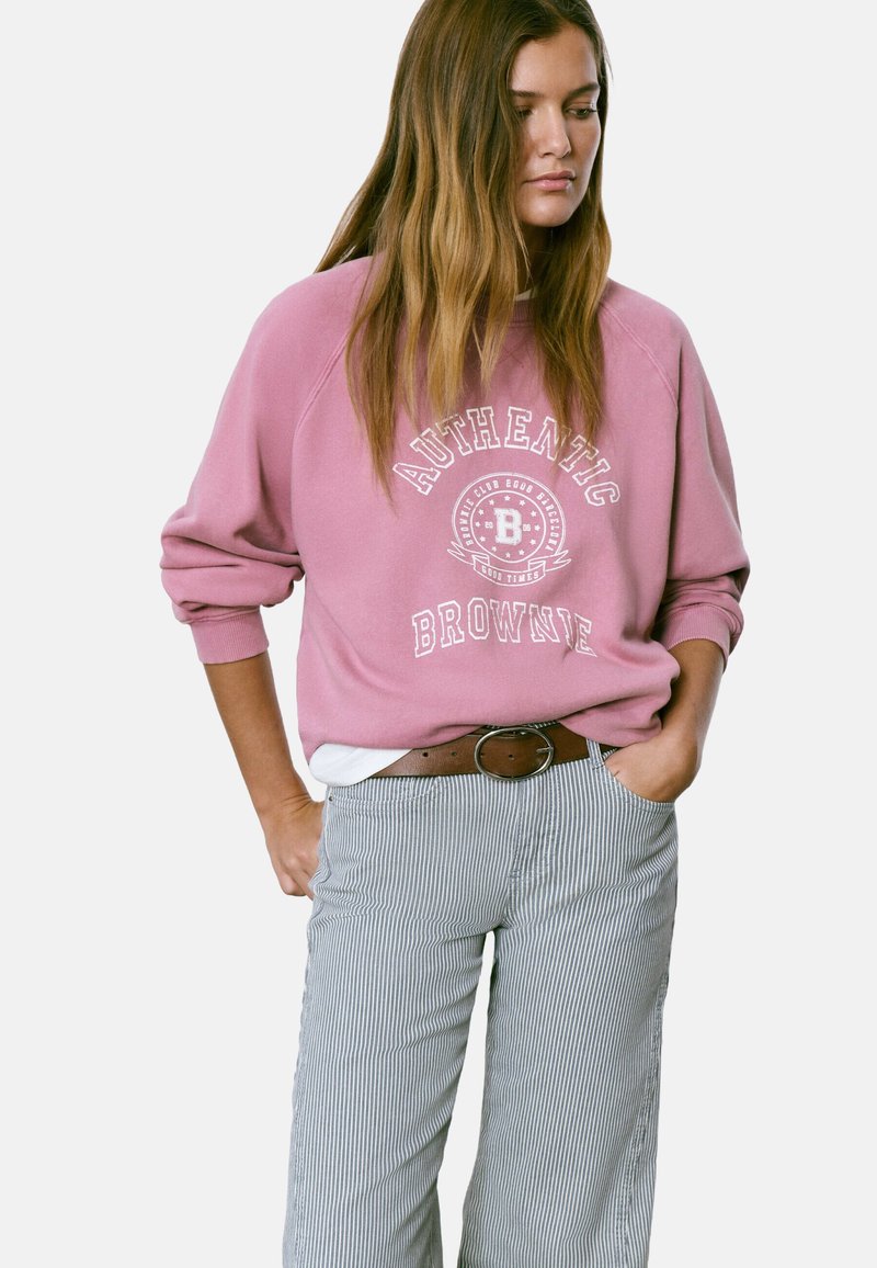 Mujer con un sudadera rosa holgada con el texto "Authentic Brownie" y pantalones a rayas, de pie con las manos en los bolsillos, mirando hacia abajo.