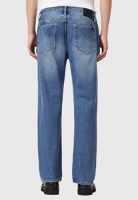 Niebieskie dżinsy o luźnym kroju, zauważalne tylne kieszenie oraz czarna metka w pasie; gładka faktura i wyblakłe wykończenie.