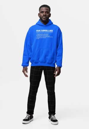 Mand iført en lyseblå hættetrøje med hvid tekst, der oplister fem ostebaserede interesser, sorte bukser med hvidt rudemønster og sorte sneakers.