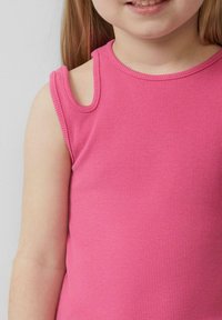 Młoda dziewczyna uśmiechająca się, ubrana w bezrękawnik różowy w prążki z małym wycięciem na ramieniu, na jednolitym tle.