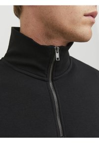Primo piano di una persona che indossa un capo zip-up nero con colletto alto, con la cerniera parzialmente chiusa.