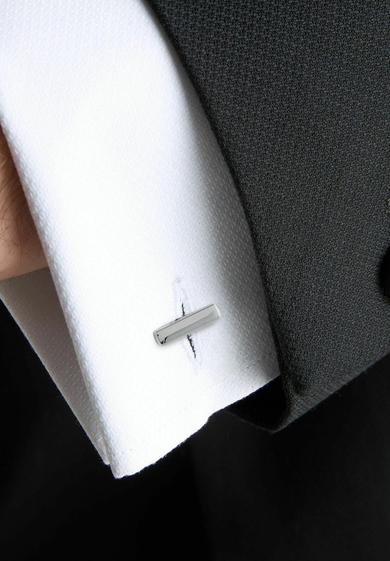 White textured cuff with a silver cufflink, secured by a black thread, against a dark grey suit fabric. Visible stitching detail.
