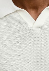 Polo en maille blanche avec une surface texturée et un col blanc uni, présentant un design à encolure en V et un motif horizontal subtil.