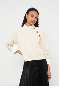 Crèmekleurige sweatshirt met drie gouden knopen op de linkerschouder, geribde zoom en opgerolde witte overhemdmanchetten, gecombineerd met een zwarte geplooide rok.