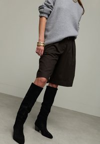 Femme portant des bottes noires jusqu'aux genoux, un short marron foncé mi-cuisse, un pull gris et des bracelets en or au poignet gauche sur fond uni.