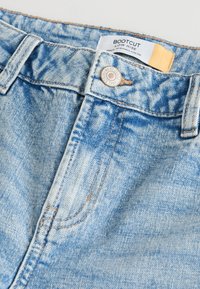 Šviesiai mėlyni džinsai su plataus apatinio siūlės stiliaus apačia, žemu liemeniu, matomais siūlais ir metaliniu mygtuku, pagaminti iš džinso su išblukusia tekstūra.