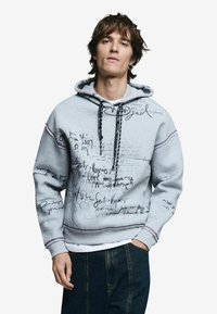 Szürke kapucnis pulóver húzózsinórral, fekete és lila kézírásszerű szöveggel és vonalakkal díszítve, kék farmernadrággal párosítva. Texturált pamut anyag.