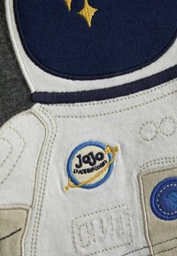 Vyšívaný dizajn astronauta na látke s námorníckym modrým, bielym a béžovým prevedením. Obsahuje logo "JoJo Space Explorer" a podrobné šitie.