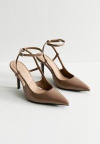Zapatos de tacón de punta afilada y tipo slingback en charol marrón brillante, con delgadas correas alrededor del tobillo y una plantilla suave de color beige.