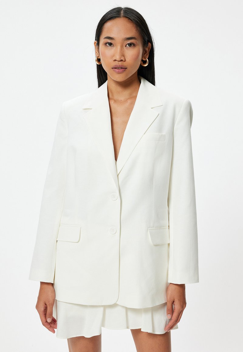 Witte op maat gemaakte blazer met een enkelvoudig ontwerp, normale revers en twee voorzakken. Zachte stof met een gladde textuur, minimale details.