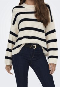 Femme portant un pull en tricot crème à rayures horizontales noires, un jean bleu foncé, et une ceinture noire à boucle dorée.