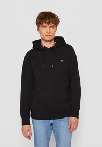 Felpa nera in cotone, con tasca frontale, cappuccio regolabile e un piccolo logo sul petto. Indossata con jeans azzurri chiari.
