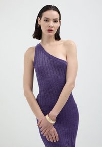 Donna con capelli corti e scuri che indossa un vestito viola a spalla unica, a coste, con un bracciale d'oro e piccoli orecchini a cerchio su uno sfondo neutro.