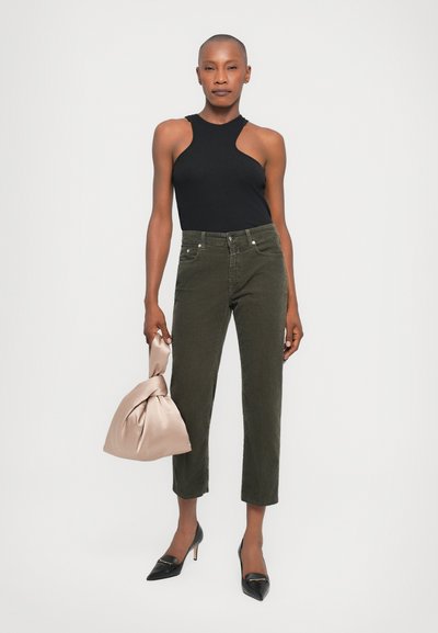 Black sleeveless top, olive green corduroy pants, and pointed black heels. Holds a knotted, light beige bag. Simple, modern design.