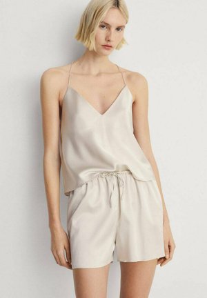 Mujer con un camisón de satén de color beige claro con tirantes finos y pantalones cortos con cordón a juego, de pie contra un fondo claro y liso.