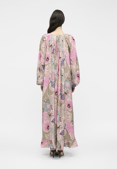 Γυναίκα με μακριά σκούρα μαλλιά που φοράει ένα μακρύ, φαρδύ φόρεμα με λουλουδένιο σχέδιο σε ροζ, μωβ και μπεζ αποχρώσεις, στεκόμενη με την πλάτη προς το φακό σε λευκό φόντο.