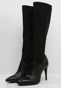 Botas negras hasta la rodilla con puntera y talón de cuero liso, caña de tela flexible, punta puntiaguda y cremallera lateral para cierre.