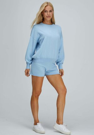 Femme blonde debout, portant un pull à manches longues bleu clair, un short assorti et des baskets blanches devant un fond gris uni.