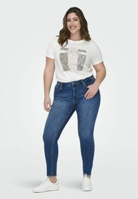 Biała koszulka z grafiką, niebieskie obcisłe dżinsy i białe trampki. Koszulka ma wzór zwierzęcy oraz tekst w czerni i różu.