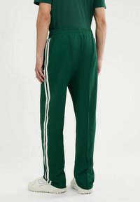 Zielone sportowe spodnie z białymi paskami po bokach, wykonane z gładkiego materiału. Posiadają elastyczny pas i luźny krój, zestawione z białymi trampkami.