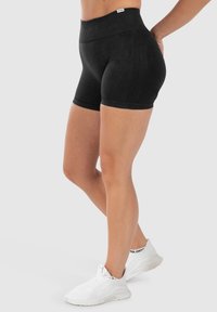 Hoch taillierte schwarze Shorts aus weichem Stoff, mit glatter Textur und figurbetontem Design, kombiniert mit weißen Sportschuhen.
