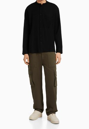 Camicia nera a maniche lunghe con colletto alla cinese e taschino frontale, abbinata a pantaloni cargo verde oliva con tasche laterali e sneakers neutre.