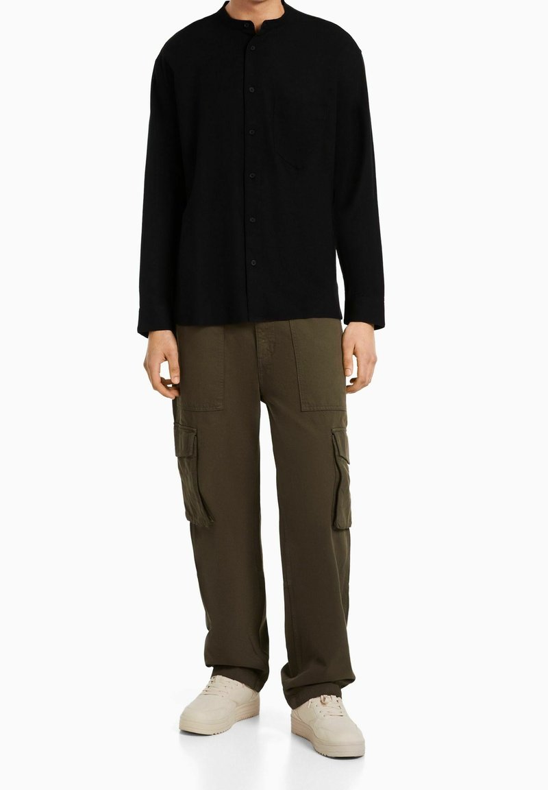 Camicia nera a maniche lunghe con colletto alla cinese e taschino frontale, abbinata a pantaloni cargo verde oliva con tasche laterali e sneakers neutre.