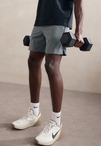 Calções atléticos cinzentos, halteres pretos, sapatilhas Nike bege com um acabamento texturizado e meias brancas com um logótipo. Fundo de superfície plana.