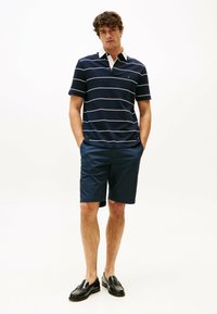 Polo a righe blu navy e bianche con colletto, abbinato a pantaloni corti blu navy. Il modello è in piedi con le mani in tasca, indossando mocassini neri.