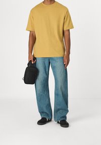 Camiseta de algodón amarilla, de manga corta y corte holgado, combinada con vaqueros anchos de mezclilla azul claro. Bolsa de tela negra en la mano. Zapatillas negras.