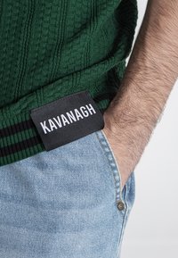 Suéter de punto texturizado verde con dobladillo a rayas en negro y verde, con una etiqueta negra que lleva el texto blanco "KAVANAGH" visible en el bolsillo.