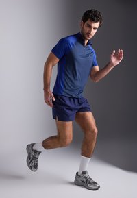 Ανδρική αθλητική ενδυμασία με ναυτικό μπλε κοντομάνικο πόλο και ναυτικό μπλε σορτσάκι. Το υλικό φαίνεται ελαφρύ. Περιλαμβάνει γκρίζα αθλητικά παπούτσια και λευκές κάλτσες.