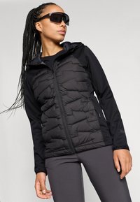 Gilet rembourré noir avec fermeture éclair, doté d'une texture matelassée et d'une capuche, associé à un haut noir à manches longues et un pantalon gris.